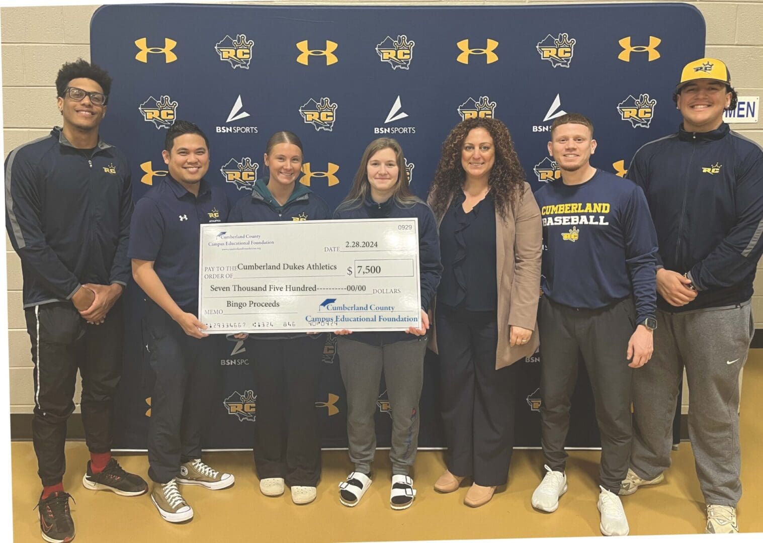 AthleticsCheckPresentation Group of student athletes and coaches presenting a large donation check to a representative, inside a gymnasium with sports banners in the background.