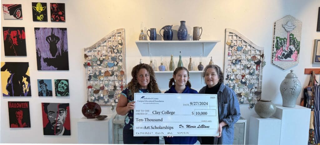 Three women holding a large check in a cozy shop filled with pottery and crafts.