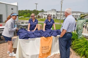 Two women in navy shirts stand behind a table with folded navy shirts, talking to an older man outdoors.